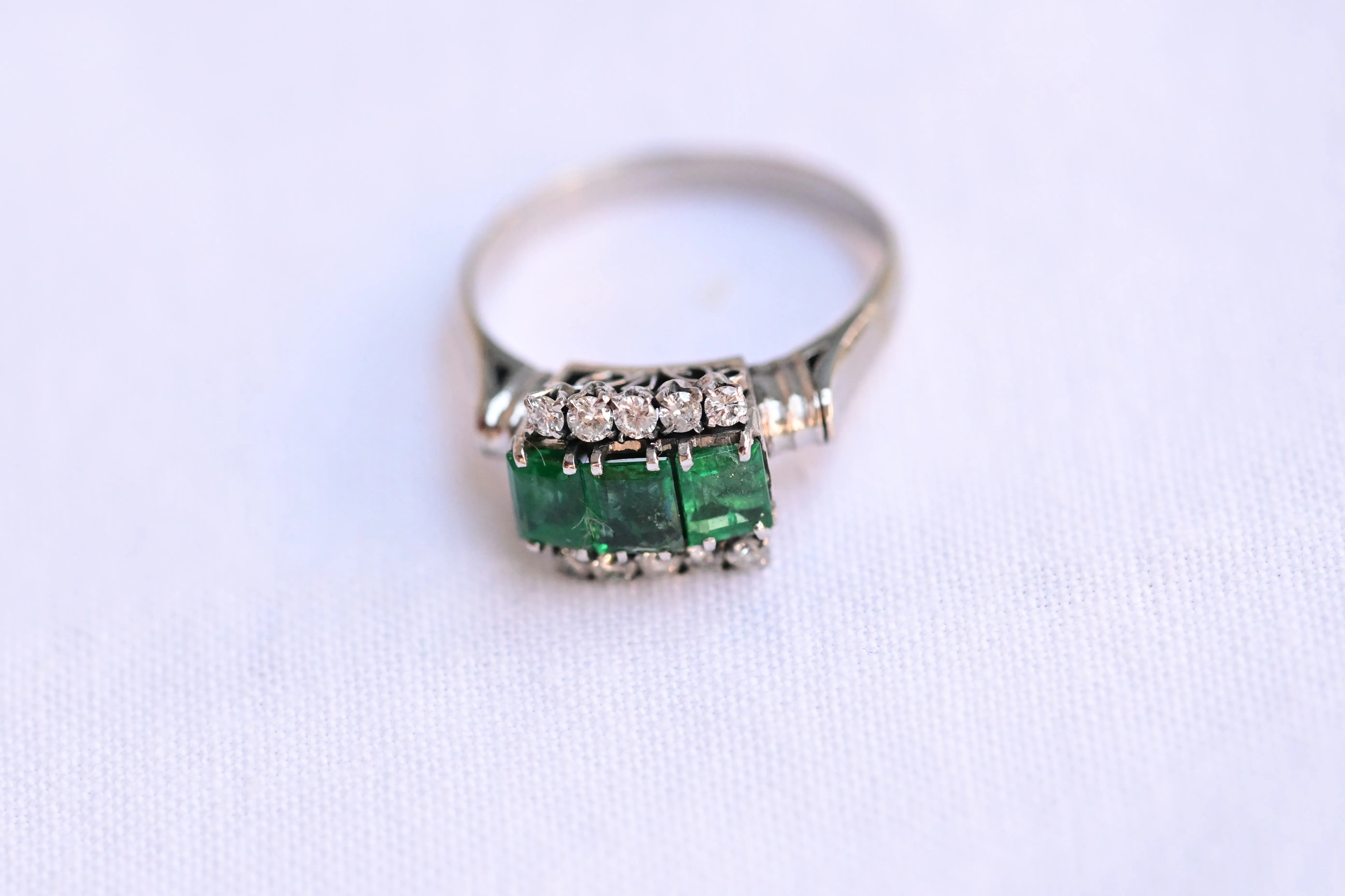 18K white gold vintage ring featuring 3 emeralds surrounded by 10 round cut diamonds.